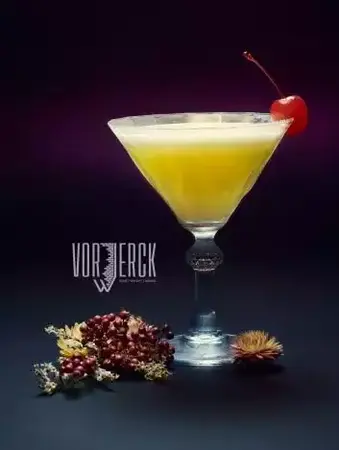 Gelber Cocktail im Martini-Glas mit Kirsche, dekoriert mit Blüten, vor dunklem Hintergrund.