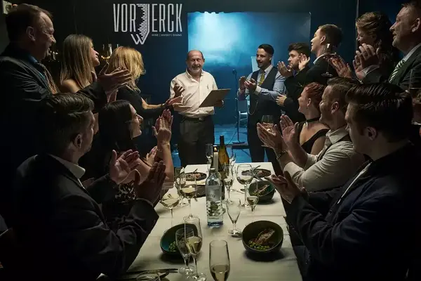 Eine festlich gekleidete Gruppe applaudiert einem Mann bei einer feierlichen Ehrung im Restaurant.