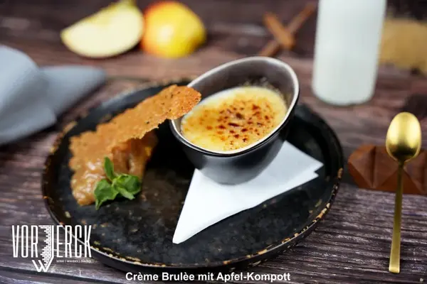Crème Brulée mit Apfel-Kompott und Karamellhippe auf rustikalem Holztisch serviert.