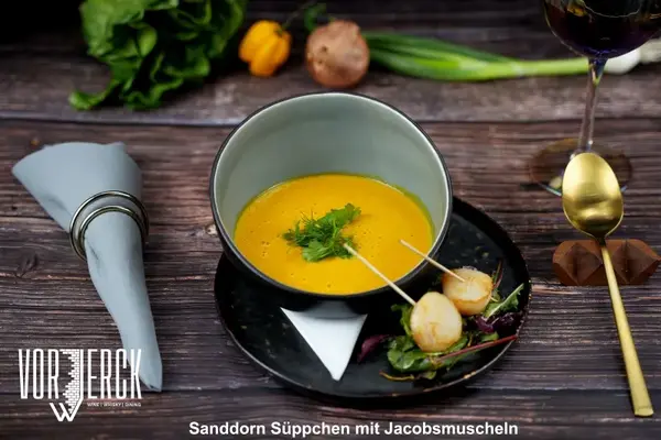 Sanddorn-Suppe mit Jakobsmuscheln und frischen Kräutern auf elegantem Holztisch serviert.