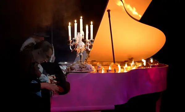 Ein brennender Flügel mit Kerzenleuchter, Pianist spielt auf Bühne im Scheinwerferlicht.