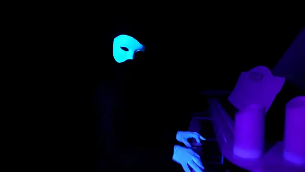 Eine Person mit leuchtender Maske spielt Klavier im Dunkeln bei Schwarzlicht-Effekt.